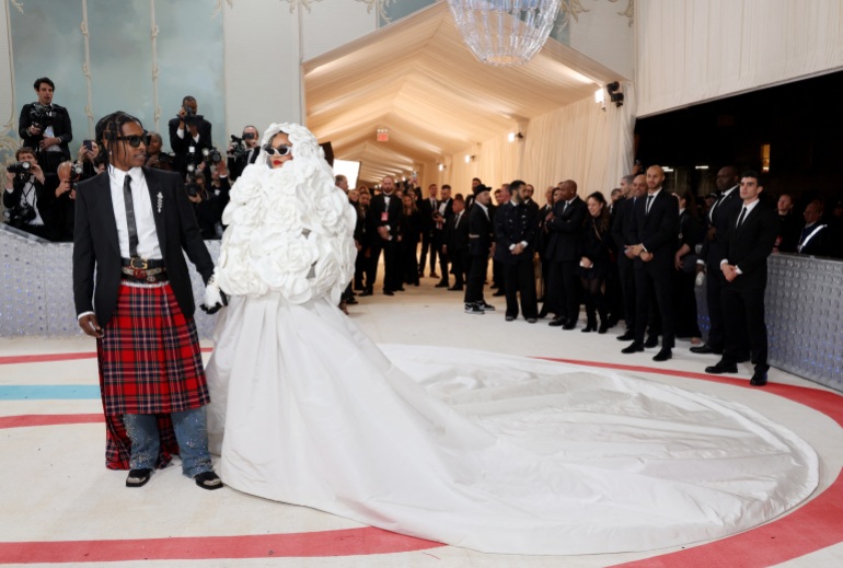 ريهانا وشريكها ASAP Rocky يقفان أمام المصورين في Met Gala.  إنها ترتدي فستانًا أبيض مع قطار طويل.  يرتدي نقبة ترتان مع قميص أبيض وسترة سوداء.