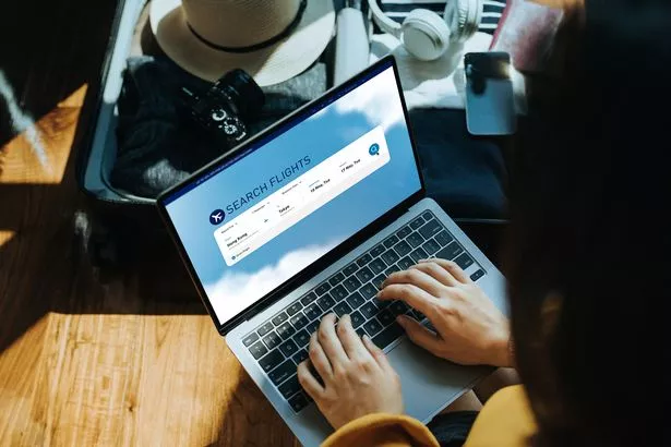 امرأة آسيوية شابة تتسوق عبر الإنترنت للحصول على تذكرة الطيران على موقع شركة طيران مع كمبيوتر محمول في المنزل ، جالسة بجانب حقيبة مع قبعة الشمس والكاميرا والملابس وسماعات الرأس والهواتف الذكية وجواز السفر. الاستعداد لقضاء الإجازة. 
