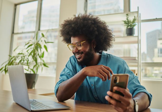 رجل أعمال إبداعي سعيد ، شاب ، مع أفرو ، يجلس على مكتب في مساحة مكاتب مريحة ، يرتدي قميصًا أزرق يحمل هاتفًا محمولًا محمولًا ويبحث عن كمبيوتر محمول.