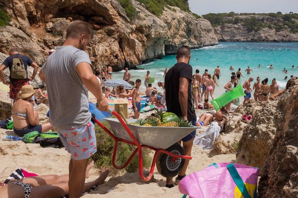Mallorca ، Balearic Isles ، إسبانيا 14 يوليو 2018 رجلان يحملان العجلات المحملة مع الأناناس الطازجة والبطيخ للبيع على شاطئ Caló des Moro المزدحم في Santanyi ، جنوب Mallorca