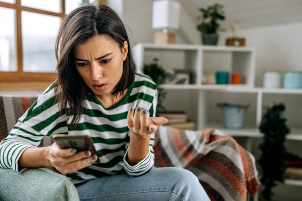 امرأة شابة محبطة ومكتئبة مع هاتف ذكي في أيديها أثناء جلوسها على الأريكة في المنزل