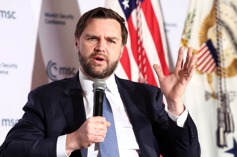 يرفع JD Vance يدًا في لفتة ويحمل ميكروفونًا مع الآخر وهو يتحدث في مؤتمر على المسرح.