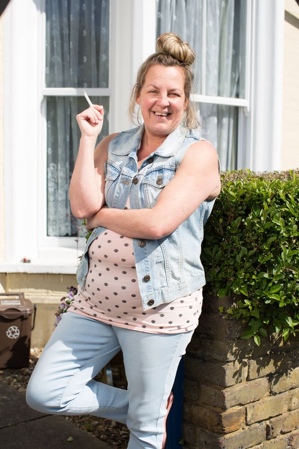 لورين ستانلي ، في قمة وردية ، سترة الدنيم والجينز ، تميل على جدار على مجموعة من EastEnders.
