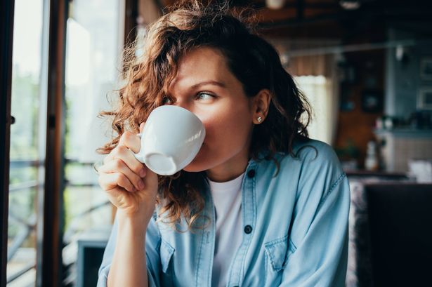 صورة لامرأة شابة جميلة سعيدة ترتدي قميصًا أزرق تشرب القهوة جالسة على طاولة في مطعم يبحث من النافذة في يوم الصيف.