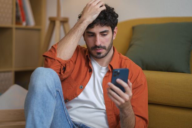 صورة لشاب ملتح جالسًا على أرضية غرفة معيشته وهو ينظر إلى الهاتف
