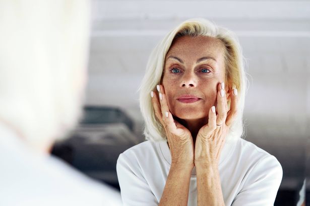 قرب من امرأة 65s الأقدم تقف أمام المرآة ، لمس وجهها برفق يديه ، وفحص الجلد ، وتقييم نتائج روتين العناية بالبشرة. ترطيب ، نظام مضاد للشيخوخة ، مفهوم الشيخوخة بأمان