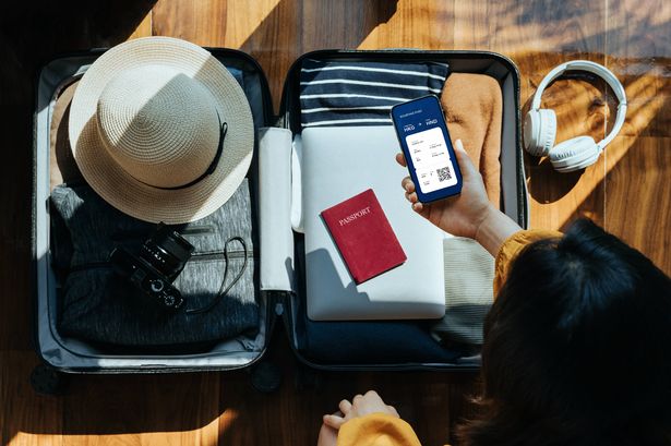 منظر علوي للمرأة الآسيوية التي تحمل هاتفًا ذكيًا تعرض تذكرة طيران إلكترونية فوق حقيبة مفتوحة مع الملابس ، وقبعة الشمس ، والكاميرا ، وسماعات الرأس ، والكمبيوتر المحمول ، وجواز السفر على أرضية خشبية ضد ضوء الشمس. ملحقات المسافر. مفهوم السفر والعطلات