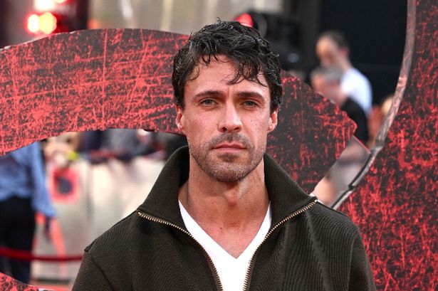 Olly Rix ، في قمة بيضاء ، سترة خضراء ، بنطلون أسود وأحذية ، على سجادة حمراء في يونيو 2025.