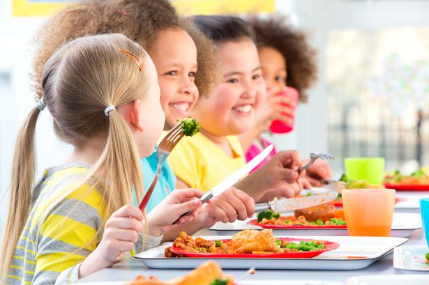 يمكن رؤية أربعة أطفال على طاولة عشاء مدرسية مع وجبات أمامهم. إنهم ينظرون إلى شيء ويبتسمون ، مع أدوات المائدة في أيديهم. طفل واحد تناول مشروب ويتطلع نحو صديقه ، الذي ينظر إليه.