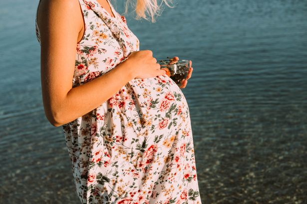امرأة حامل ذات شعر أبيض على الشاطئ في ثوب ونظارات شمسية عند غروب الشمس