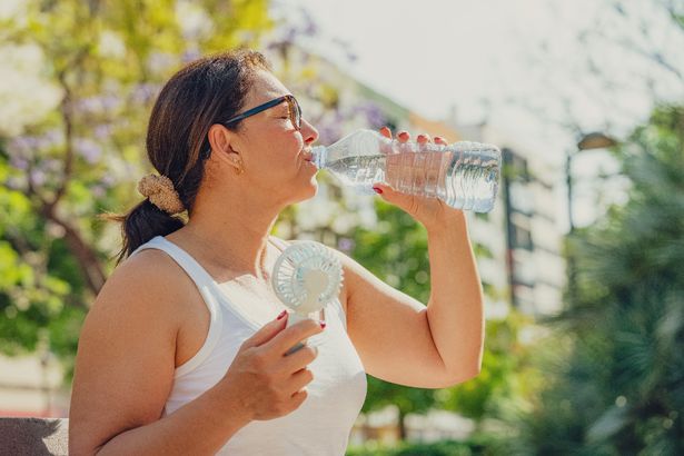 المرأة تعاني من ارتفاع درجة حرارة مياه الشرب واستخدام مروحة اليد الكهربائية المحمولة في الهواء الطلق. موجة الحرارة