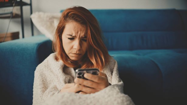 تظهر امرأة قوقازية للبالغين في منتصفها أثناء جلوسها على أرضية غرفة المعيشة ، ملفوفة في سترة مريحة ، تسعى إلى الاتصال أو الهاء على هاتفها الذكي.