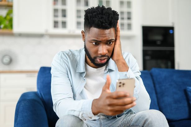 تلقى الشاب الشاب المزعج الذي يحمل الهاتف الذكي ، وينظر مع تعبير خيبة الأمل في شاشة الهاتف ، تعليقًا سيئًا في الشبكات الاجتماعية من Hater ، أثناء الجلوس على الأريكة