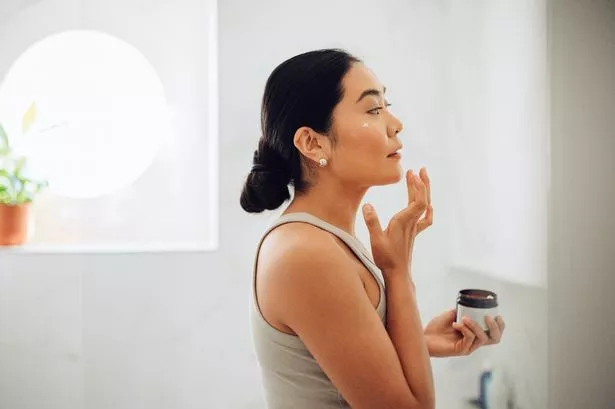 امرأة آسيوية تقف في حمامها ، وتمسك كريم الوجه وتضعه على خديها ووجهها.