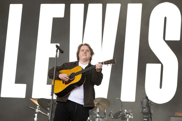 لويس كابالدي ، في قمة بيضاء ، سترة داكنة وسروال ، يحمل غيتارًا على خشبة المسرح في Glastonbury 2025.