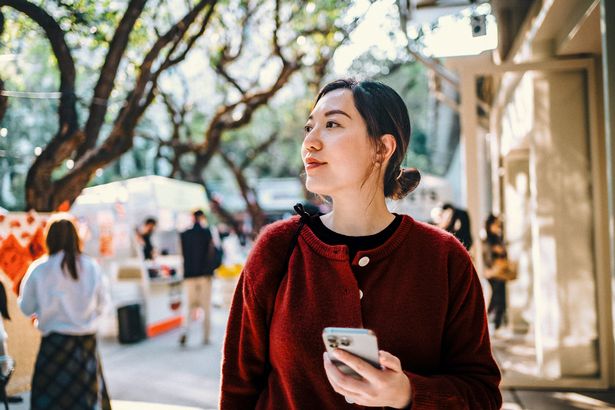 امرأة آسيوية شابة تستخدم الهاتف الذكي أثناء الاستمتاع بالمشي واستكشاف المدينة في سوق الشوارع. إنها تشرب المعالم السياحية وتتجول على طول الشارع مع المشاعر الصينية الجديدة.