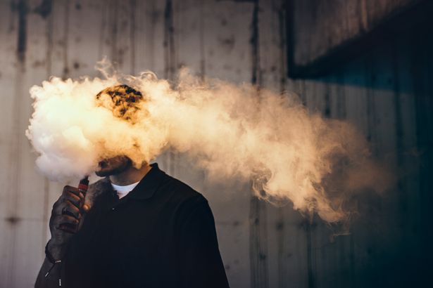 صورة تباين مظلمة وعالية لذكور شاب لها سحابة من بخار النيكوتين "دخان" يتدفق من فمه. تدخين vape ، أو "vaping" ينمو في شعبية ، وكذلك الوقوع في ظل لوائح الدولة والحكومة أكثر صرامة. صورة أفقية مع مساحة النسخ.