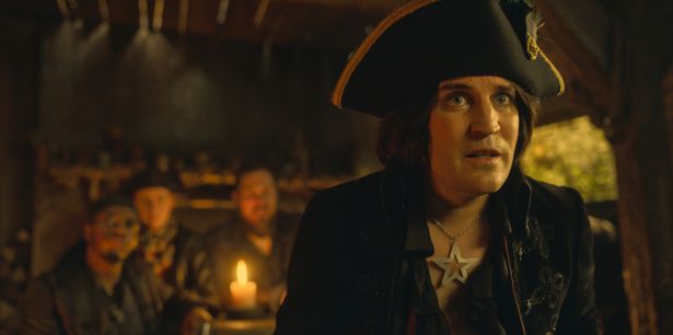 Noel Fielding في ملابس سوداء ، بما في ذلك قبعة سوداء وقلادة نجم فضية ، في مشهد من مغامرات Dick Turpin بالكامل بالكامل