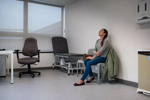 منظر واسع الطول على نطاق واسع لامرأة متوسطة البالغات جالسة في غرفة فحص الأطباء في انتظار رؤيتها ، فهي تمسك بذراعها وتنظر وتشعر بالقلق. ترتدي ملابس غير رسمية في المستشفى في نيوكاسل ، إنجلترا.