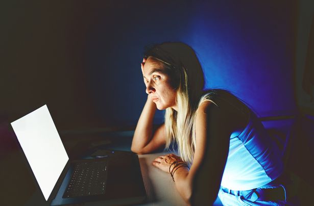 امرأة شابة تنظر إلى جهاز الكمبيوتر المحمول في غرفة مضاءة بشكل خافت