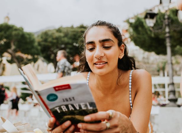 امرأة هندية جميلة تقع على بطنها على شاطئ مزدحم. تقرأ كتاب دليل السفر.