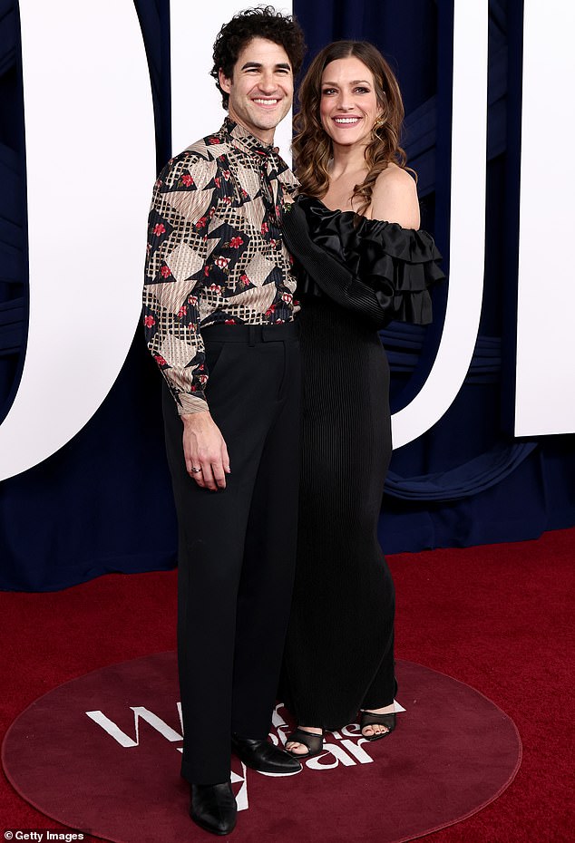 دارين كريس، يرتدي بلوزة ذات طباعة هندسية داخل مجموعة من السراويل السوداء عالية الخصر، وهو يقف بجانب زوجته ميا سوير