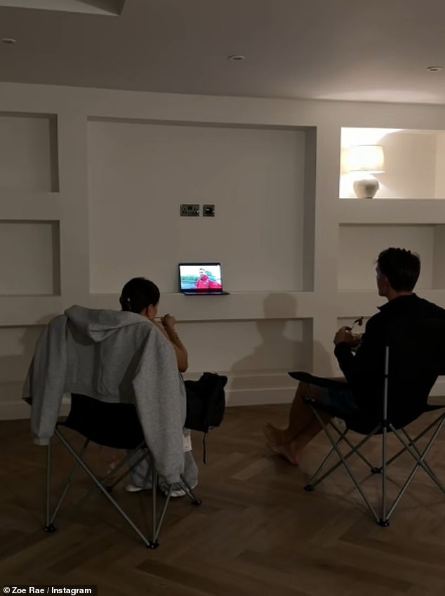 وفي صورة أخرى، يسترخي الزوجان، اللذان ليس لديهما حاليًا أي أثاث في الطابق السفلي، على كرسيين للاستلقاء أثناء مشاهدة التلفزيون على جهاز كمبيوتر محمول في غرفة المعيشة الجديدة الخاصة بهما.