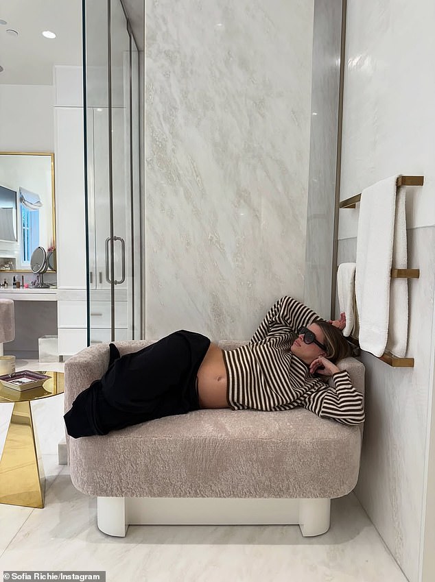 استراحت على أريكة مريحة باللون البيج أمام الحمام بينما كانت ترتدي بنطال صالة أسود