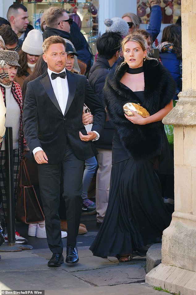 يصل اثنان من الحاضرين يرتديان اللون الأسود بالكامل، وترتدي المرأة شالًا رقيقًا وتحمل حقيبة مطلية بالذهب